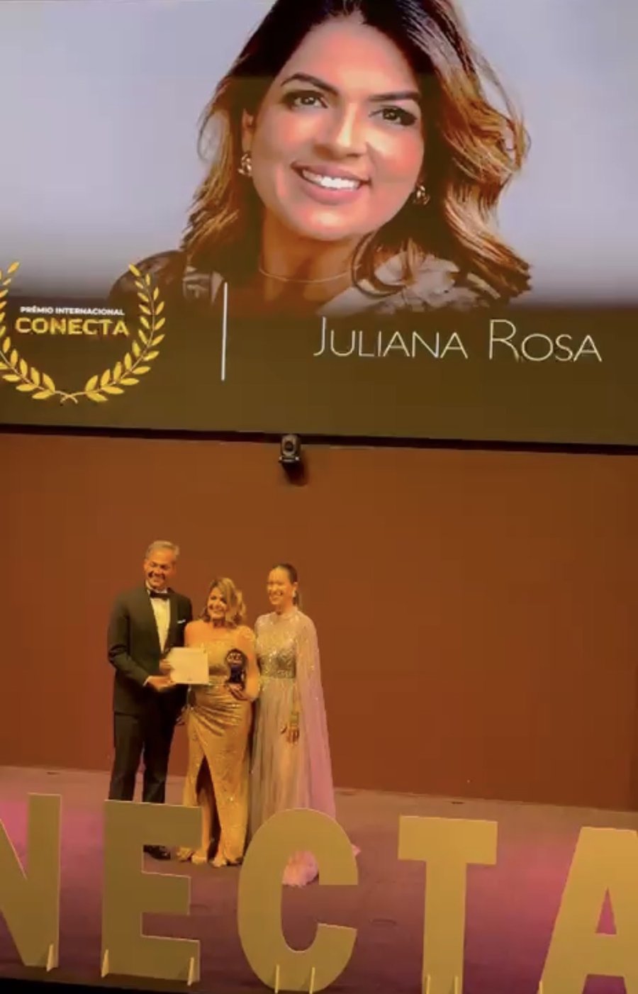 Juliana Rosa recebendo o Prêmio Internacional Conecta no palco, com seu nome no telão